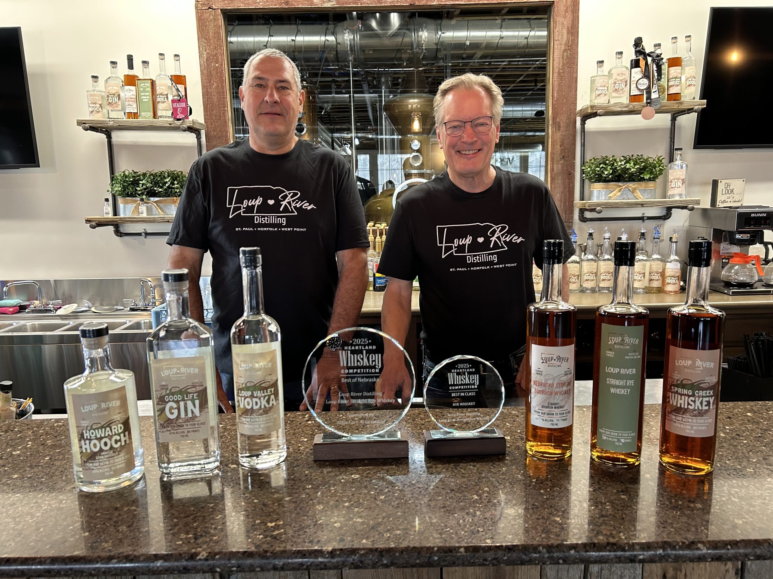 Eric Montemagni and Dirk Petersen are standing behind a distillery bar with a display of Loup River Distilling spirits and award trophies, showcasing Nebraska-made whiskey, gin, vodka, and hooch.