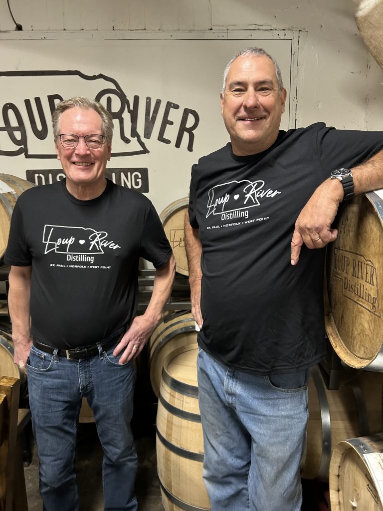 Dirk Petersen and Eric Montemagni are standing in a distillery surrounded by wooden whiskey barrels, wearing Loup River Distilling T-shirts and smiling in front of a branded wall.